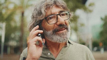 Friendly middle aged man with gray hair and beard wearing casual clothes using his mobile phone. Mature gentleman in eyeglasses talking on cell phone outdoors
