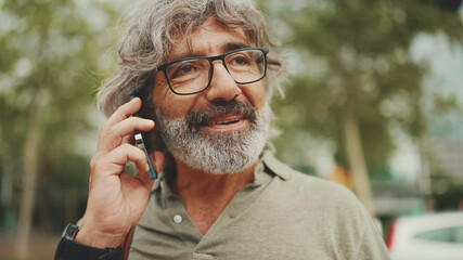 Friendly middle aged man with gray hair and beard wearing casual clothes using his mobile phone. Mature gentleman in eyeglasses talking on cell phone outdoors