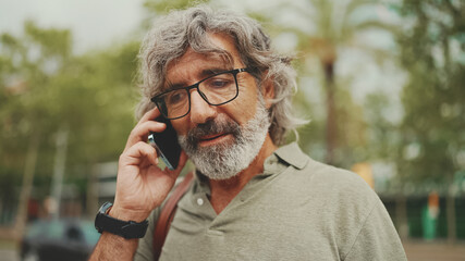 Friendly middle aged man with gray hair and beard wearing casual clothes using his mobile phone. Mature gentleman in eyeglasses talking on cell phone outdoors - Powered by Adobe