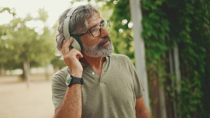 Friendly middle-aged man with gray hair and beard wearing casual clothes listening to music on headphones. Mature gentleman in eyeglasses enjoys music outdoors