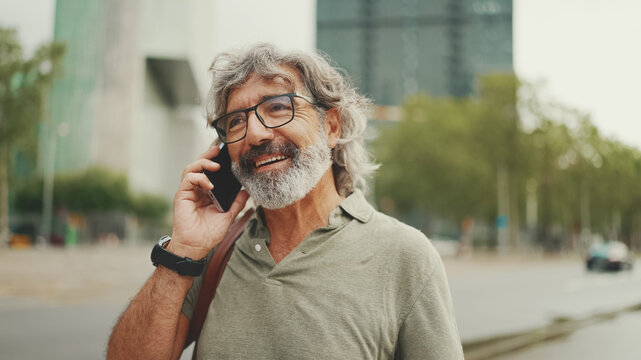 Friendly middle aged man with gray hair and beard wearing casual clothes using his mobile phone. Mature gentleman in eyeglasses talking on cell phone outdoors