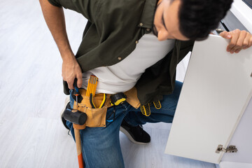 High angle view of arabian man taking screwdriver from tool belt near kitchen cabinet