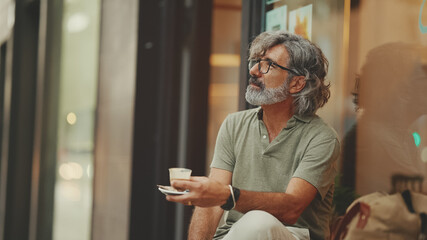 Clouse-up, thoughtful middle-aged man with gray hair and beard, wearing casual clothes, sits in street cafe. Mature gentleman in eyeglasses drinks aroma coffee