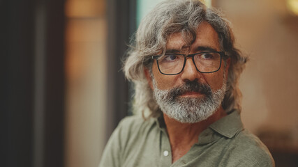 Thoughtful middle-aged man with gray hair and beard, wearing casual clothes, sits in street cafe. Mature gentleman in eyeglasses