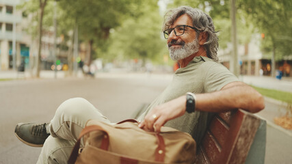 Positive middle-aged man with gray hair and beard wearing casual clothes sits on bench. Mature...
