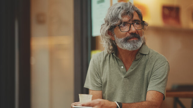 Clouse-up, thoughtful middle-aged man with gray hair and beard, wearing casual clothes, sits in street cafe. Mature gentleman in eyeglasses drinks aroma coffee