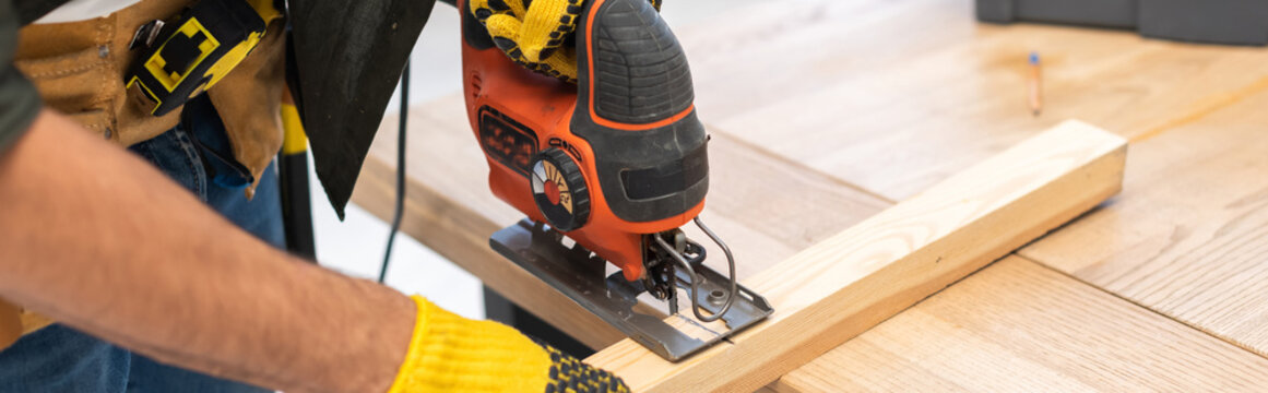 Cropped view of man in gloves using jigsaw machine on wooden plank at home, banner