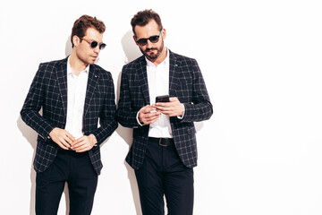 Two handsome confident stylish hipster lambersexual models. Sexy modern men dressed in black same elegant suit. Fashion male posing in studio near white wall. Shows something at his mobile phone