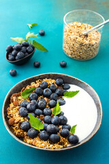 Homemade granola with yogurt, blueberries and mint leaves in a bowl on a turquoise background, delicious healthy breakfast