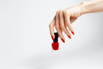 Close-up of female hand with red manicure holding a glitter nail polish of red color on white...