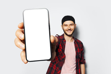Studio portrait of young smiling man holding big smartphone with blank on screen in hand, showing close to camera a device with mockup on white background. Wearing red plaid t-shirt and striped shirt.