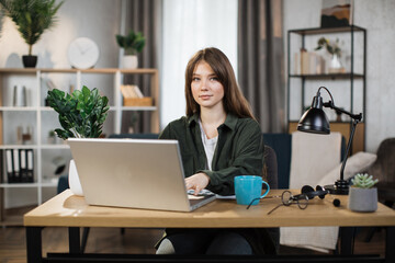 Beautiful caucasian woman with long dark hair sitting at desk and typing on laptop. Focused female in dark shirt and jeans using portable computer for work at bright office