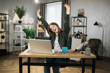 Happy caucasian woman student in casual wear sitting at table with modern laptop and keeping arms raised. Concept of people, technology and distance learning and success.