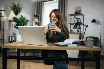 Portrait of positive caucasian female holding cup with coffee and watching favorite movie on modern laptop. Young excited woman sitting at workplace with portable computer and document.