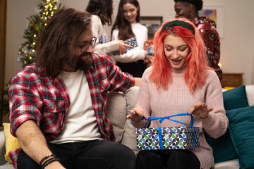 A cheerful girl with pink hair opens a gift she received from a bearded man with glasses. The excited woman slowly unties the ribbon.