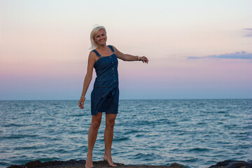 A beautiful blonde girl stands at sunset on the seashore of the ocean in a stylish denim dress, selective focus, travel vacation