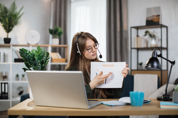 Caucasian woman presenting financial report to colleagues through video call on laptop. Female freelancer sitting at workplace and talking in headset.