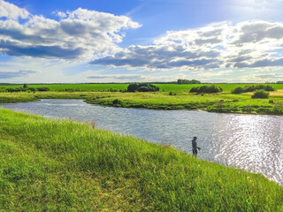 Summer landscape of wild nature, water body and silhouette of a fisherman with a fishing rod, green meadow, trees, blue sky with clouds. Clear day, open spaces, shades of summer, fresh greenery, field