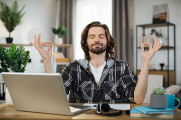 Handsome man freelancer sitting at table with closed eyes and relieving stress by meditation at...
