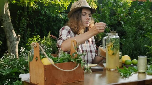 Young woman makes summer homemade lemonade cocktail in back yard garden. Woman adds brown sugar to lemonade glass jug.