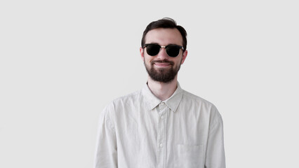 Close-up portrait of a handsome smiling young man with beard in sun glasses. Joyful student or freelancer isolated on gray background studio shot
