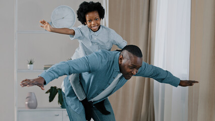 Mature african american grandpa plays with little granddaughter joyful kid pretends to be flying on plane sitting on grandfather's back funny male spread arms showing wings happy family enjoy playtime