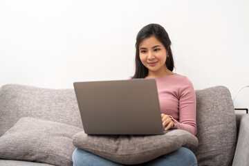 Naklejka premium cheerful asian woman using laptop while sitting on sofa in living room