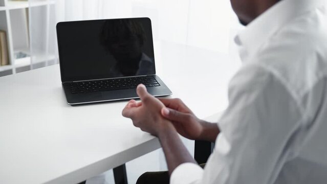 Online conference. Greeting man. Virtual connection. Unrecognizable business black guy sitting work desk with opened laptop dark screen clapping hands light room interior.