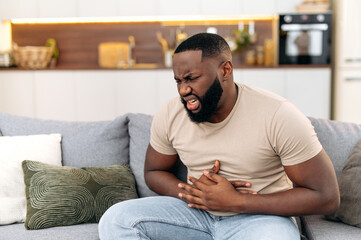 Unhappy guy of african american ethnicity, sits on a sofa in the living room, holds his hands on his stomach, grimaces from pain in his stomach, suffers from poisoning, spasm, stomach problems