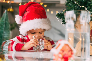 Boy toddler in a red Santa hat drinking filtered water from a glass in the kitchen. Holidays, health concept.