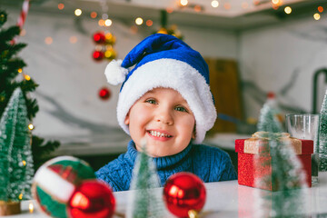 Funny toddler boy in a Santa blue hat is sitting in the kitchen with a decor for Christmas. Holidays, Traditions, baby food and health concept.