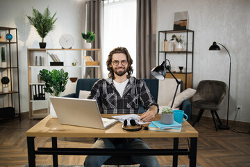 Bearded young man in checkered shirt and eyeglasses sitting at table with modern laptop and writing notes in modern office. Concept of freelance and technology.