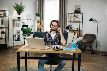 Bearded young man in checkered shirt and eyeglasses sitting at table with modern laptop and writing notes in modern office. Concept of freelance and technology.