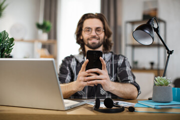 Confident young man writing message during mobile conversation at office. Caucasian male worker with long dark hair sitting at table with opened laptop.