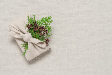 Winter gift wrapped in fabric with green thuja branch, beige linen textile background. Furoshiki...