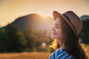 Young beautiful happy cheerful woman in a hat enjoying the sunset in a meadow with mountains view