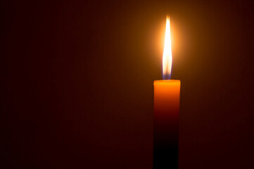 A single burning candle flame or light glowing on a yellow candle isolated on dark red or brown background on table in church for Christmas, funeral and memorial service