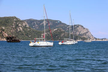 Fototapeta na wymiar Yachts with lowered sails and flags of Turkey in blue bay of Mediterranean sea on green mountains background in summer