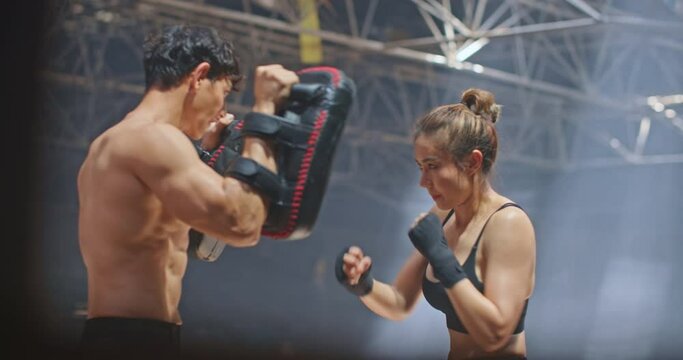 Professional female boxer doing boxing workout punching her instructor trainer in abandoned building dark hall gym. Young asian woman fighter practicing punches training with coach before competition.
