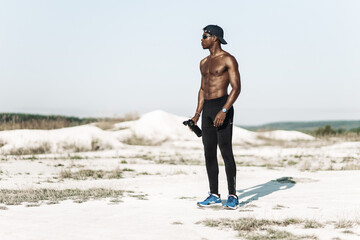 African American man with toned body holding bottle of refreshing water relaxing after workout...