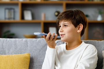 Young cute boy recording voice message on smartphone. Smiling teenager using virtual assistant app...