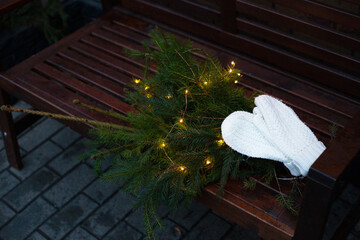 Branches of a coniferous tree decorated with a garland lie on a bench along with knitted white mittens.