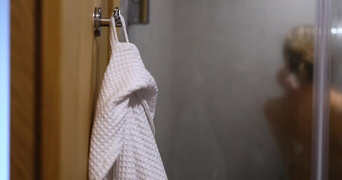 White robe on hook and naked woman taking relaxing shower in morning or evening