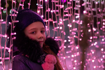 happy smiling girl in a knitted hat and scarf stands at decorative construction with a festive colorful illumination on Christmas night