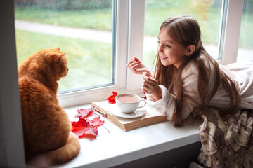 Little girl in warm terry robe lying on window sill and reading a book. Winter weekend with cat at...