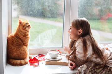 Little girl with red cat lying on window sill , reading a book and drinking cocoa. Autumn weekend with cat at home.