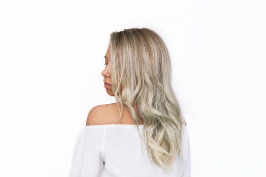 A young charming blonde woman with wavy blond hair in a white blouse standing with her back isolated on a white background. Result of coloring, highlighting, perming. Beauty and fashion