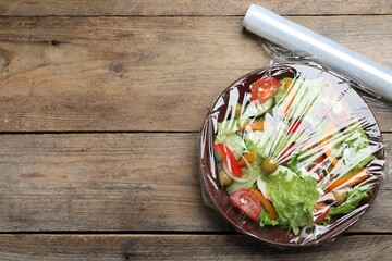 Bowl of fresh salad with plastic food wrap on wooden table, flat lay. Space for text