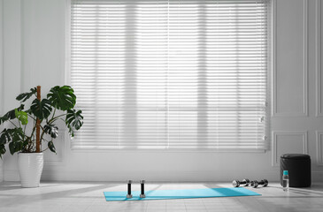 Exercise mat, dumbbells and bottle of water near window in spacious room