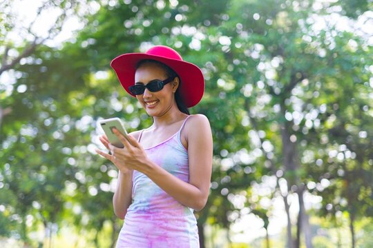 Portrait photo of the beautiful moment of a young asian beautiful lady with red hat and sunglass happily using her phone at a garden park strolling